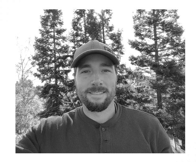A man in a baseball cap and sweater standing next to trees.